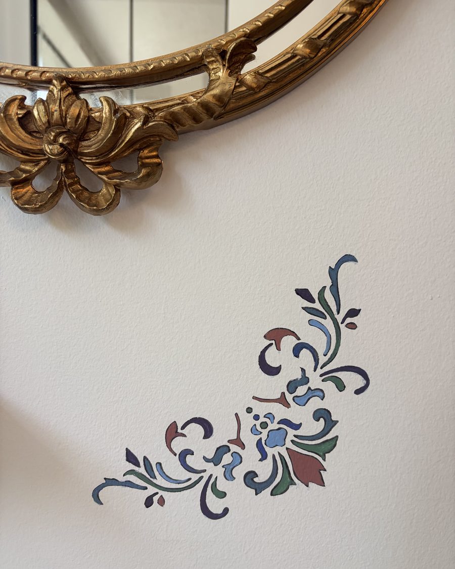 Corner of a carved gilt mirror above a hand-painted floral wall stencil in blue, green and rust on a plaster wall.