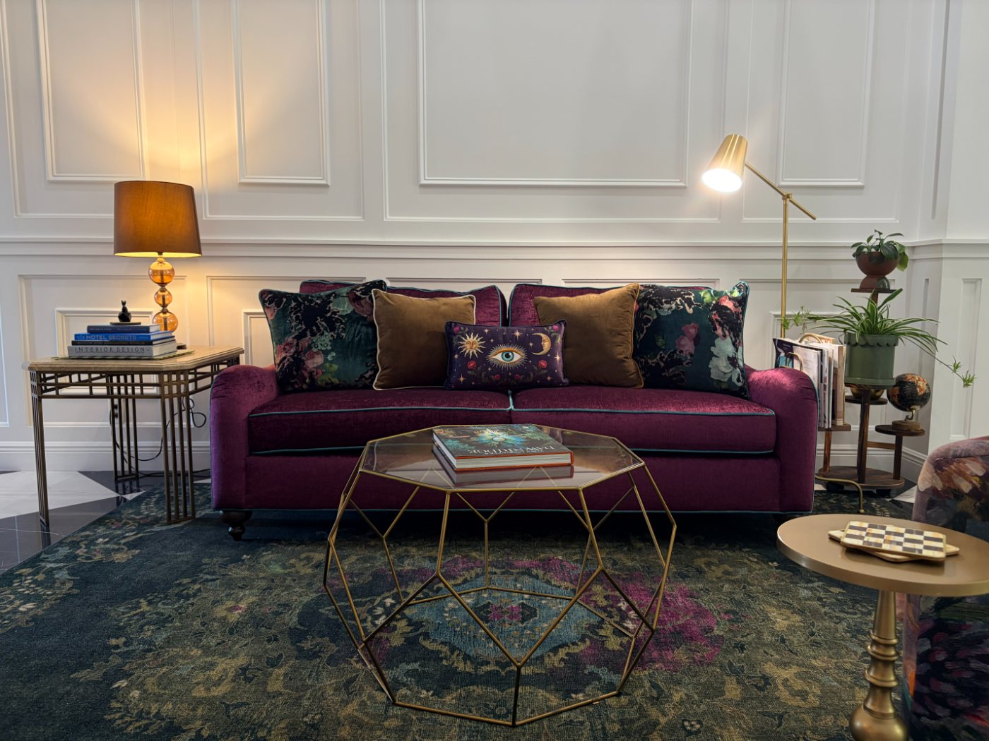 A plum velvet sofa against white panelled walls, with floral and velvet cushions around a central eye cushion, a brass geometric coffee table and a dark patterned rug.