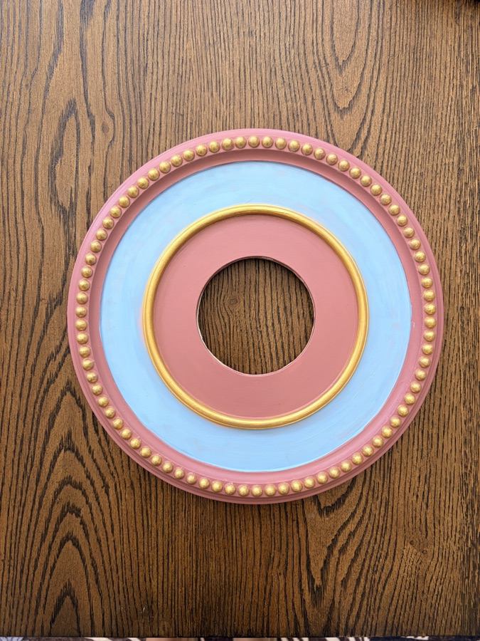 Pink and blue hand-painted ceiling medallion with gold beaded trim, photographed flat on a wooden table.