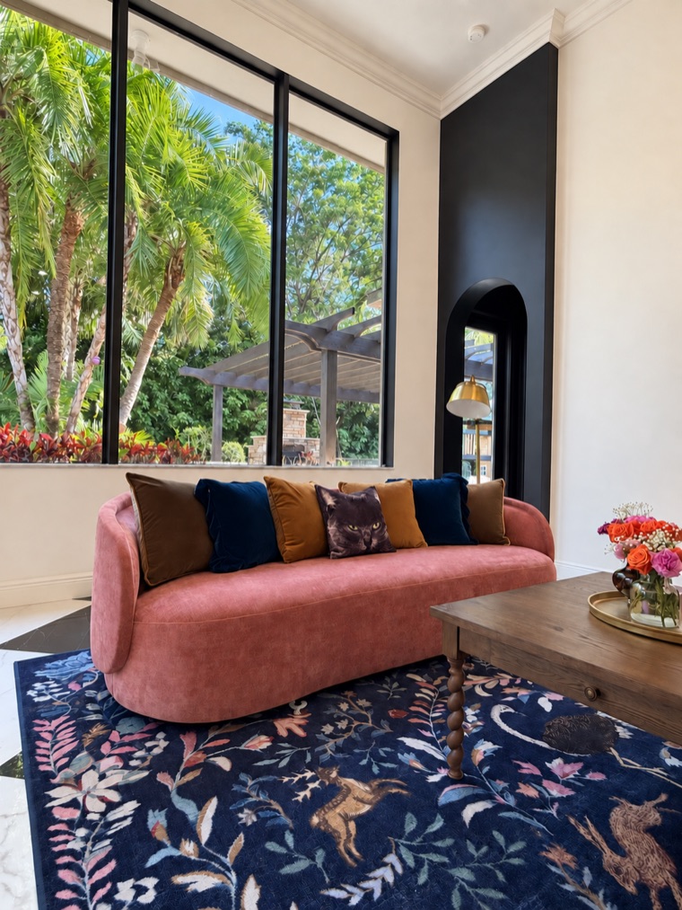 Family room with a curved rose-coloured sofa beneath a tall black-framed window, layered with velvet cushions over a deep blue botanical rug.