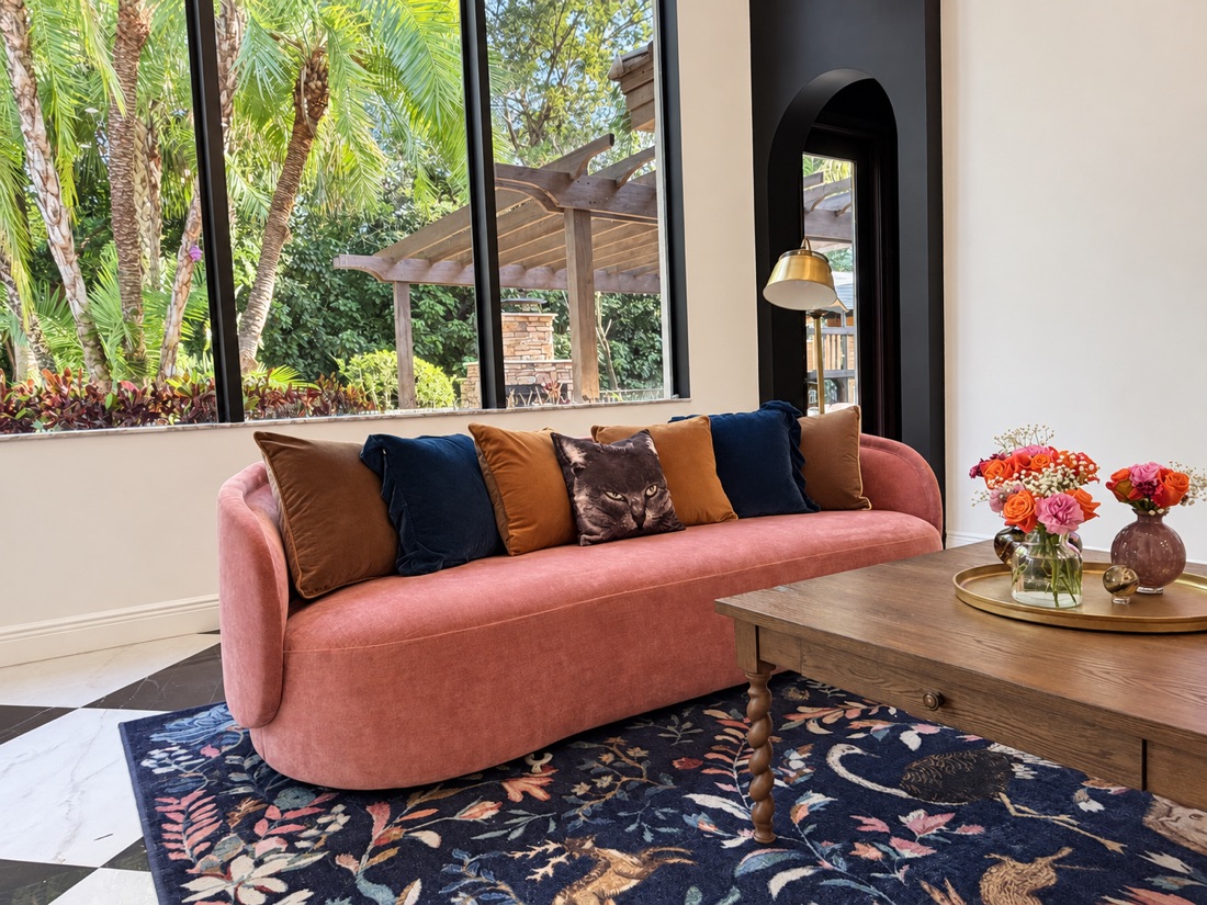 Wide view of the curved rose-coloured sofa with velvet cushions beneath tall black-framed windows onto palms, beside a timber coffee table and flowers.
