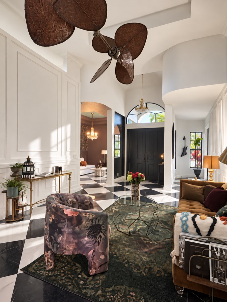 Double-height foyer with white panelling, a palm-leaf ceiling fan and a black-and-white marble checkerboard floor, with an arched opening through to the kitchen.