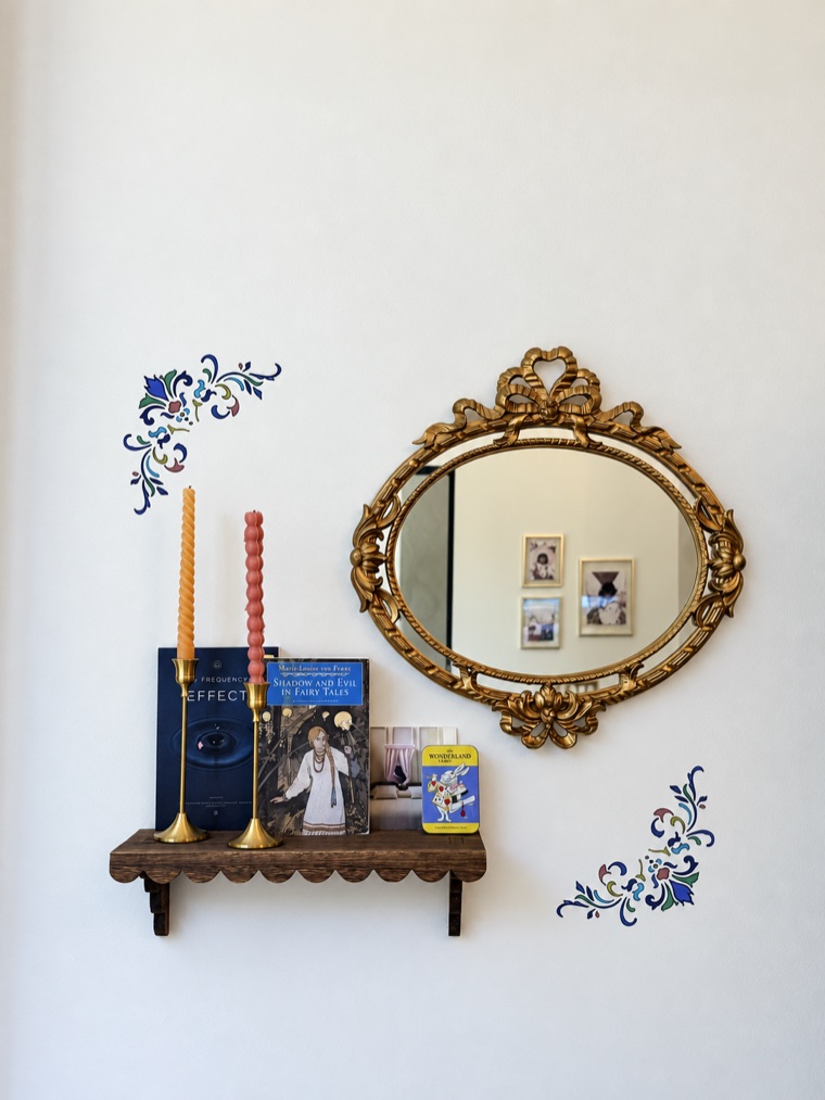 Carved gilt mirror above a small wall shelf with twisted candles and books, framed by hand-painted multicolour floral stencils on a white wall.