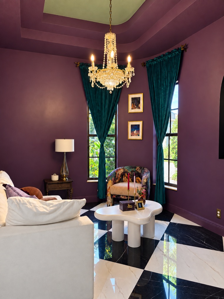 Plum-walled room under a sage-green tray ceiling, with a crystal chandelier, green velvet curtains, a white sofa and a white sculptural table on the checkerboard marble floor.