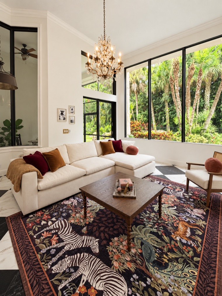 White sectional sofa with rust and burgundy cushions beneath double-height black-framed windows onto the garden, over a patterned rug, with a crystal chandelier above.