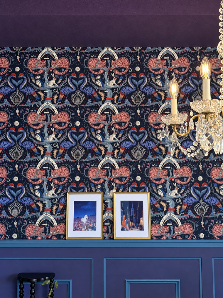 Coral, indigo and cream patterned wallcovering above plum panelling, with two framed prints and the arm of a crystal chandelier.